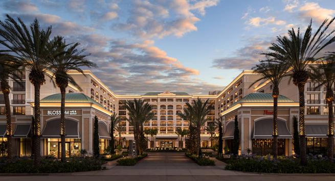 Elegant multi-story building with palm trees and storefronts, lit warmly at dusk against a partly cloudy sky.
